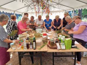 Na załączonym zdjęciu widzimy grupę kobiet, które stoją wokół stołu i przygotowują mieszanki ziołowe. Na stole Widzimy zioła, sole i wiele innych produktów ziołowych. 